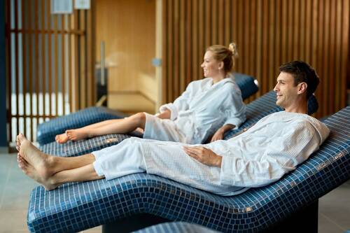 Ein Pärchen im Wellnessbereich, beide liegen im weißen Bademantel auf zwei getrennten Ruheliegen aus Stein und lächeln in die Ferne.
